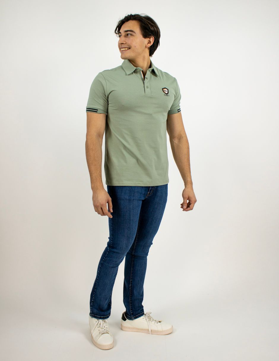 Hombre de pie con polo verde claro, vaqueros azules y zapatillas blancas sobre un fondo blanco liso.