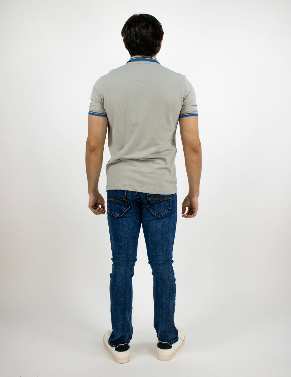 Hombre de pie de espaldas a la cámara, con una camisa polo gris, vaqueros azules y zapatillas deportivas sobre un fondo blanco.