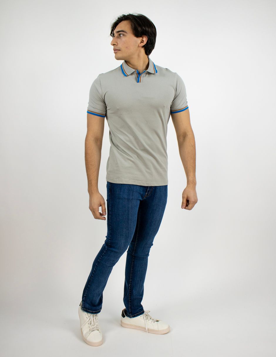 Hombre de pie con polo gris, vaqueros azules y zapatillas blancas sobre un fondo blanco liso.