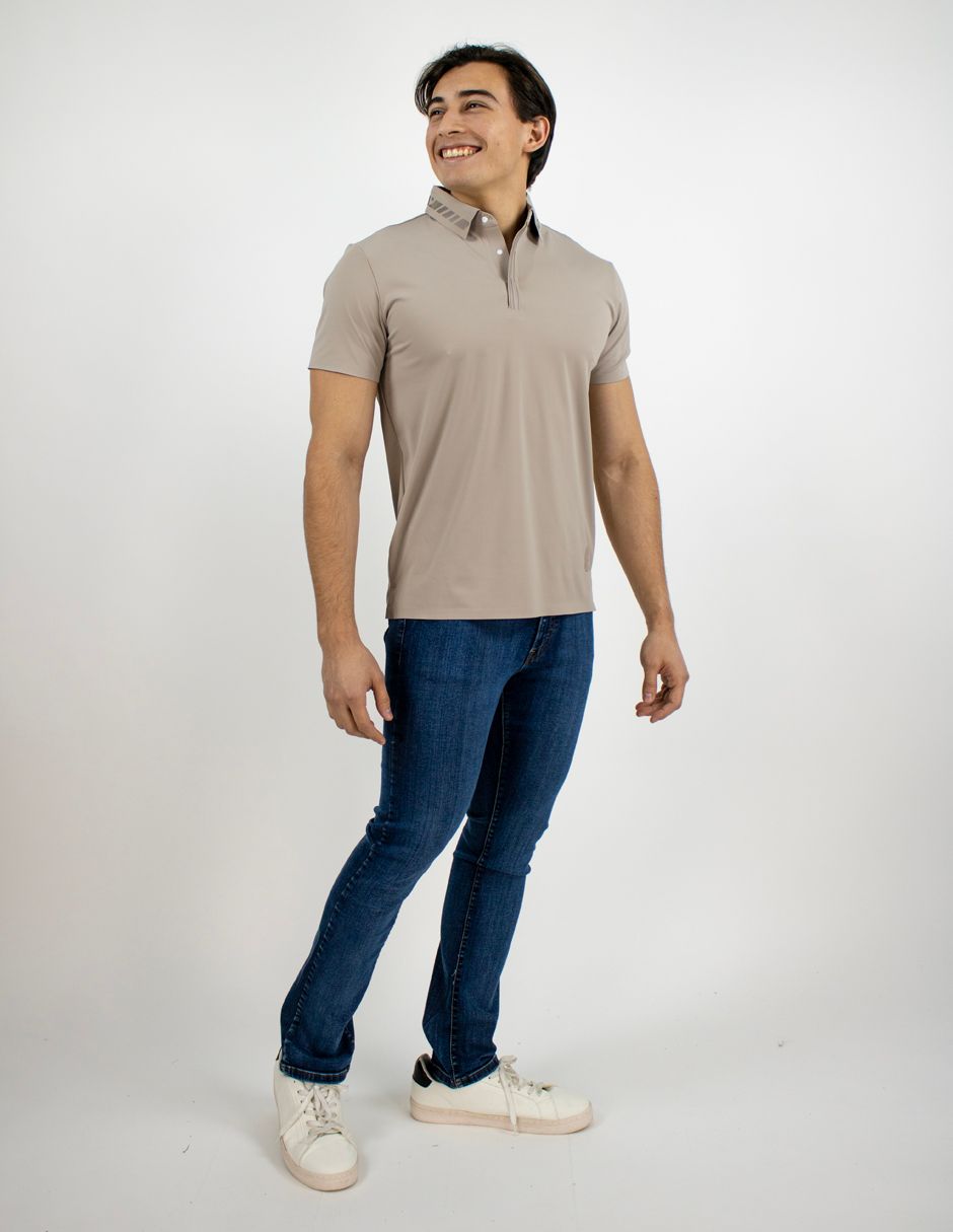 Persona de pie sonriendo con una camisa polo beige, vaqueros azules y zapatillas blancas sobre un fondo blanco liso.