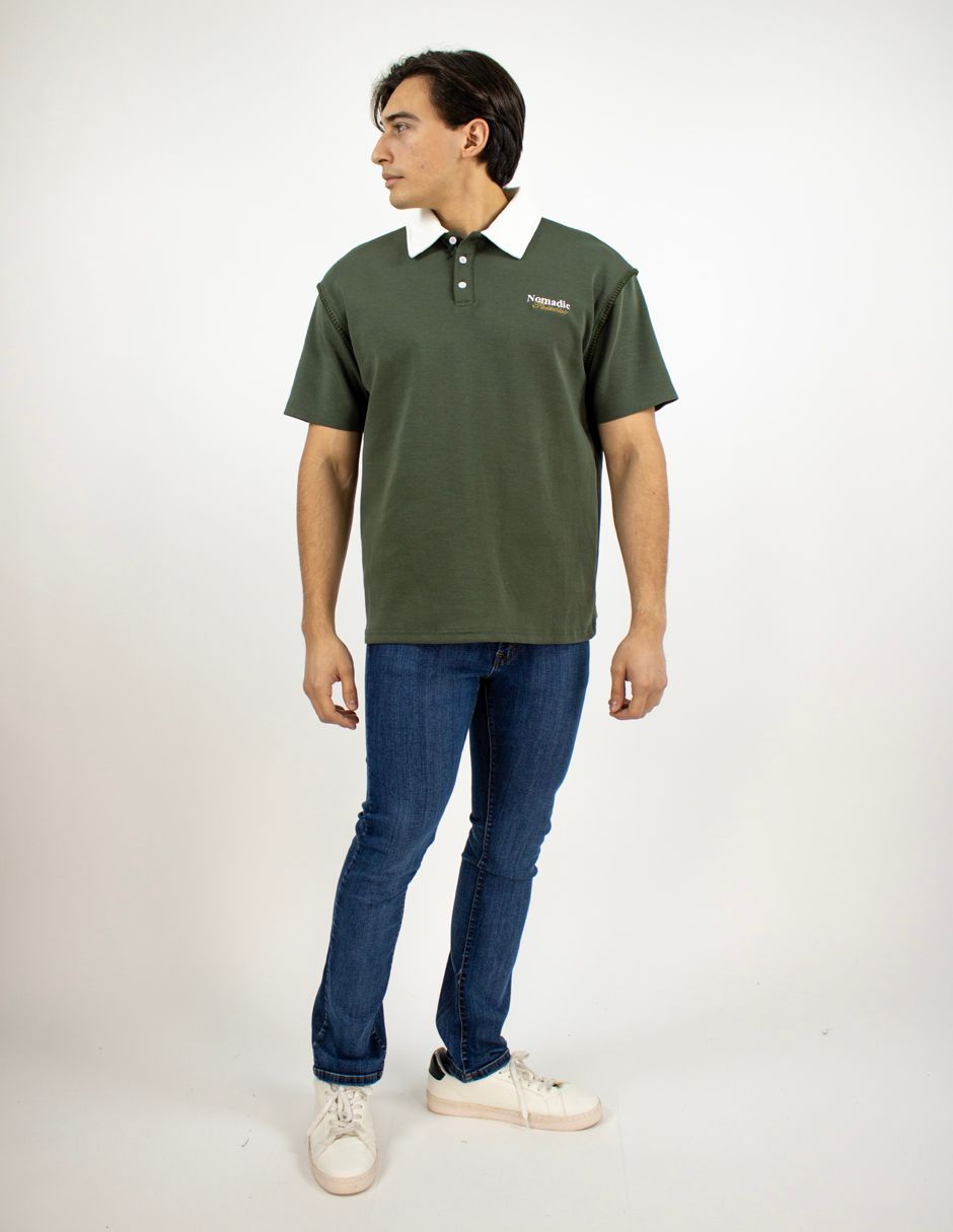 Hombre con polo verde oscuro, vaqueros azules y zapatillas blancas, de pie sobre un fondo blanco.