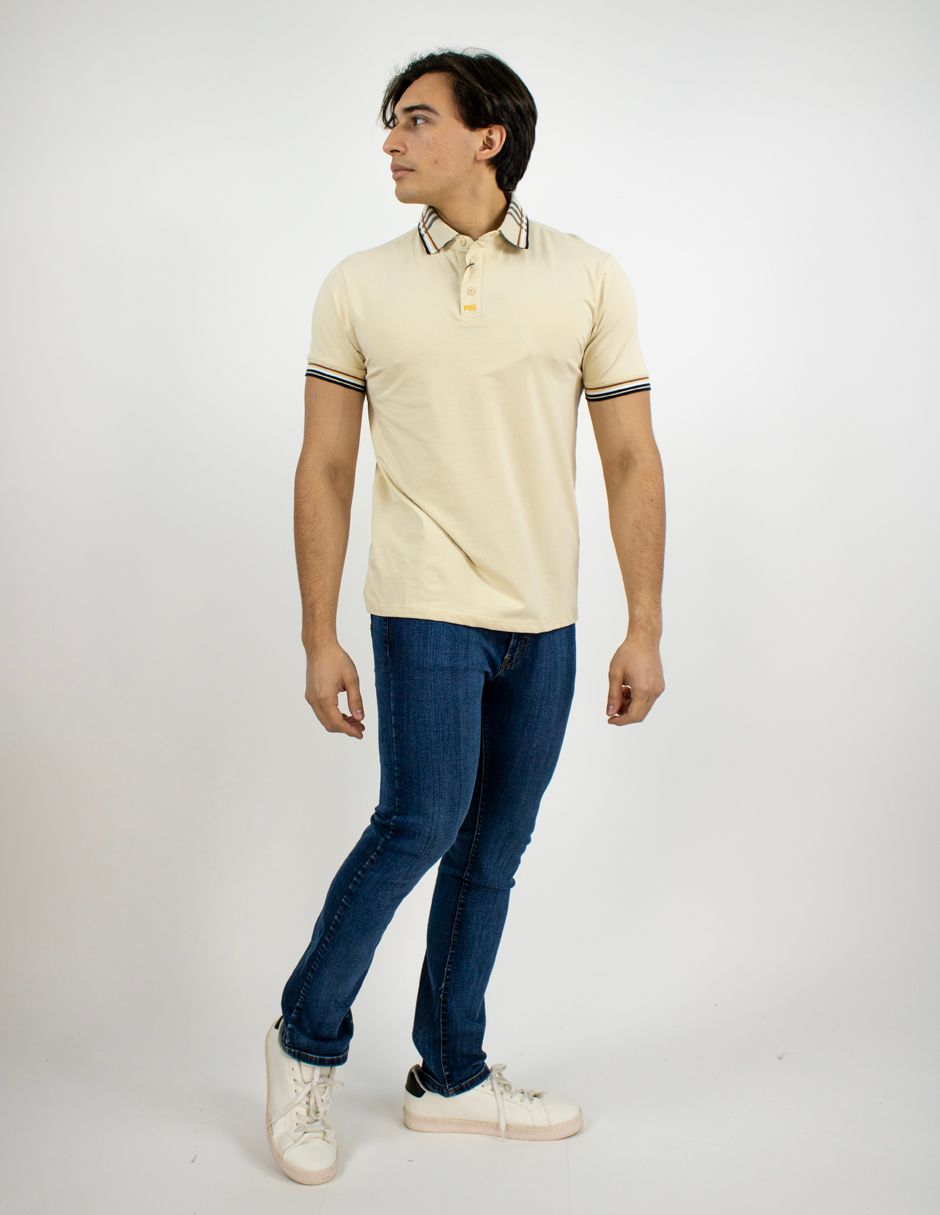 Persona de pie con polo beige, vaqueros azules y zapatillas blancas sobre un fondo blanco liso.