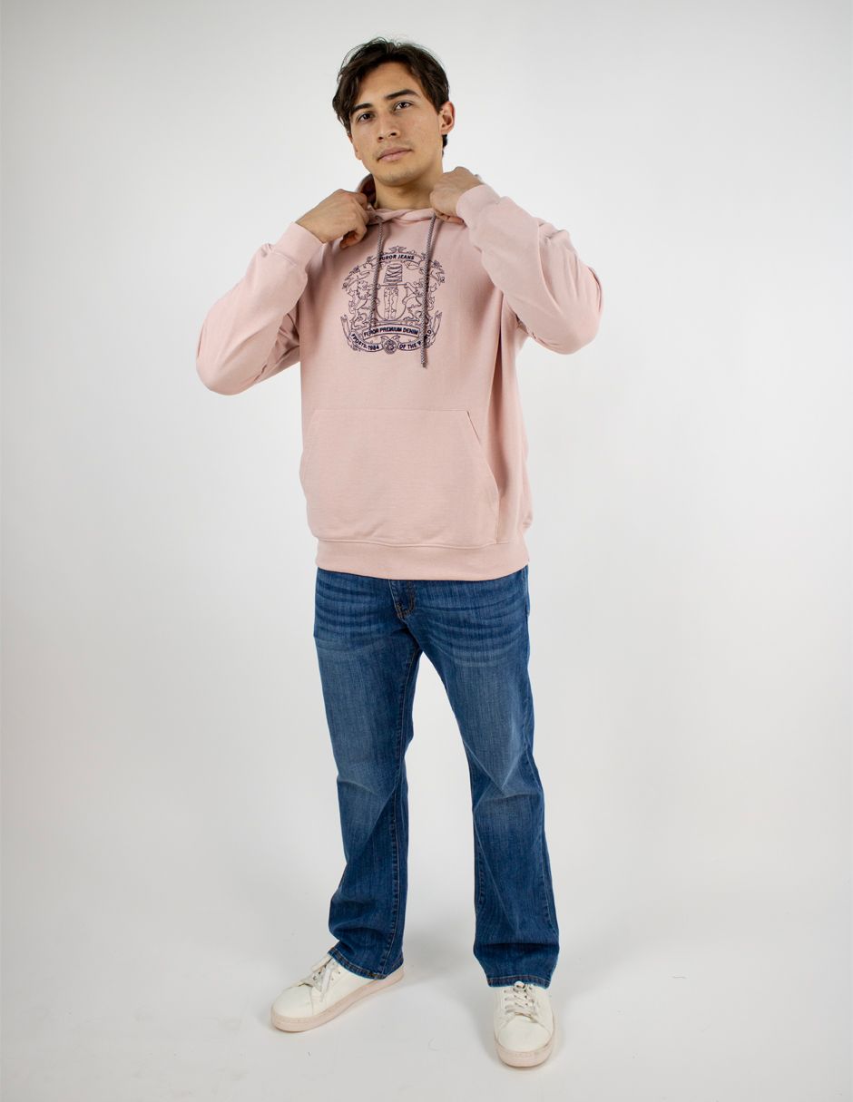 Persona de pie sobre un fondo blanco, vestida con una sudadera rosa con capucha, vaqueros azules y zapatillas blancas.