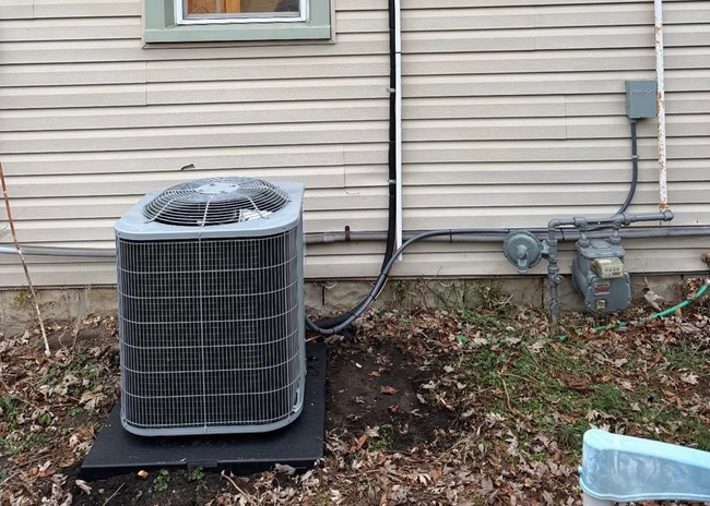 Outdoor air conditioning unit beside a house, with leaves and utility lines along the siding.