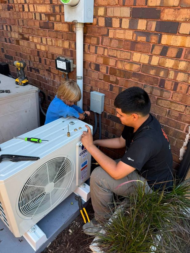 Two technicians repairing an outdoor HVAC unit beside a brick wall