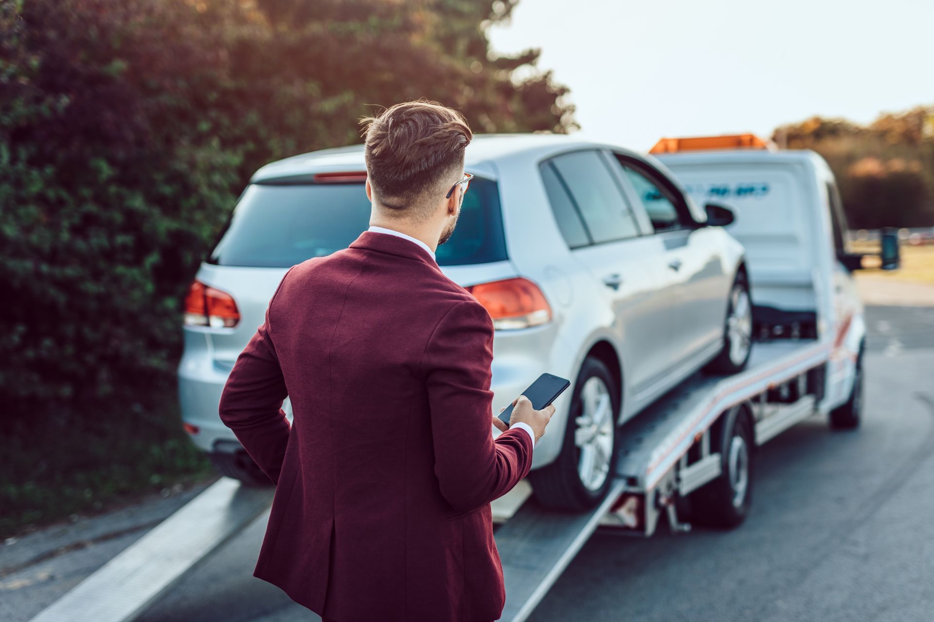Vehicle owner using a smartphone while a car is secured on a flatbed tow truck for transport.
