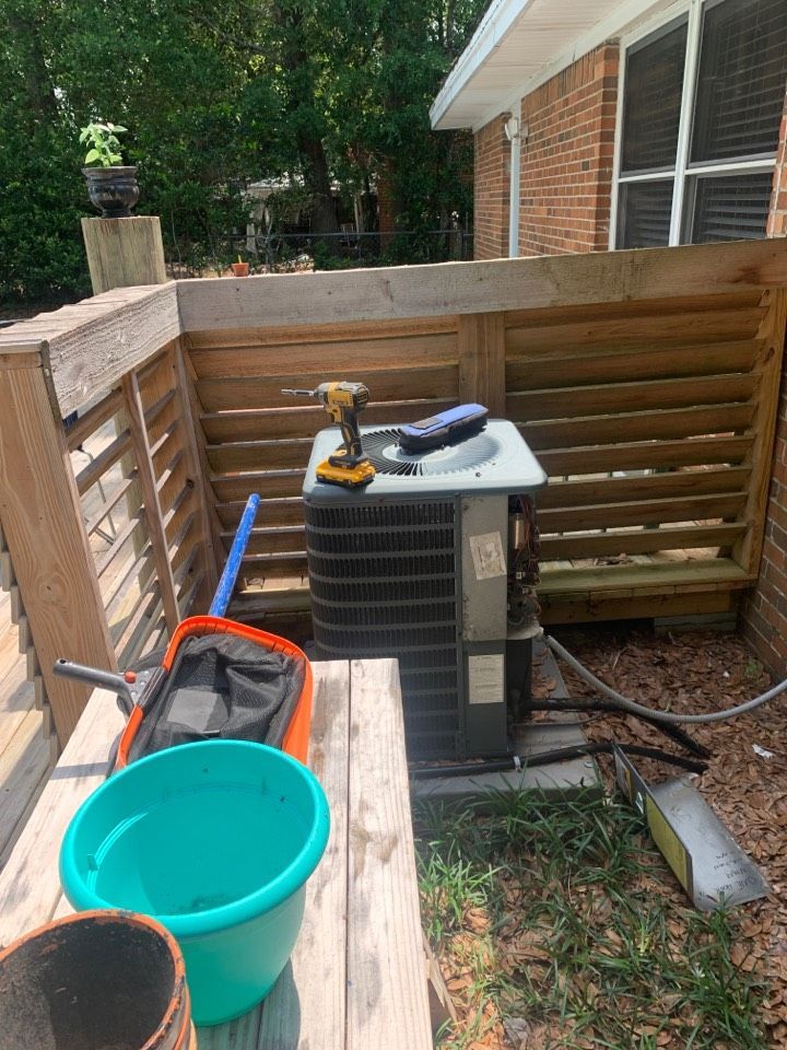 An outdoor AC unit sits behind a wooden privacy fence, with a drill, tools, and a teal bucket on a nearby wooden surface.