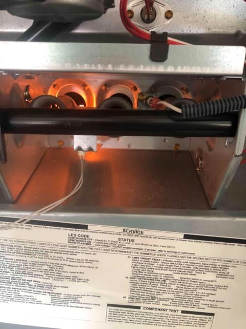 Internal view of a residential furnace with a glowing orange hot surface igniter and burner tubes above a service sticker.