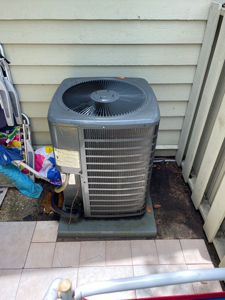 A gray outdoor air conditioning condenser unit sits on a concrete pad next to a house wall with beige siding.