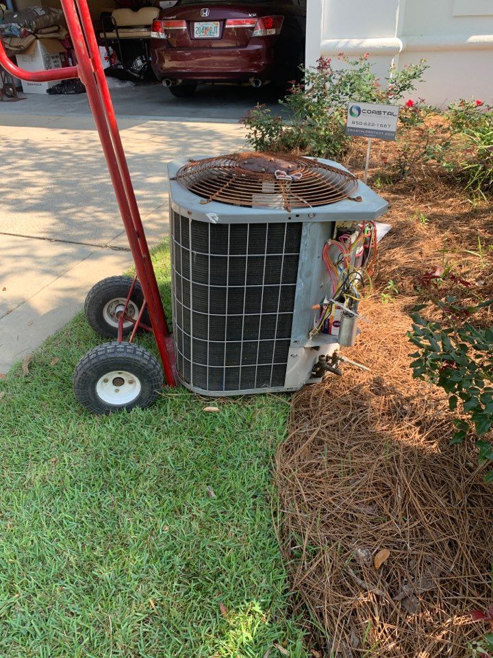 A red hand truck moves a removed grey air conditioning unit across a lawn next to a garden bed and parked vehicle.