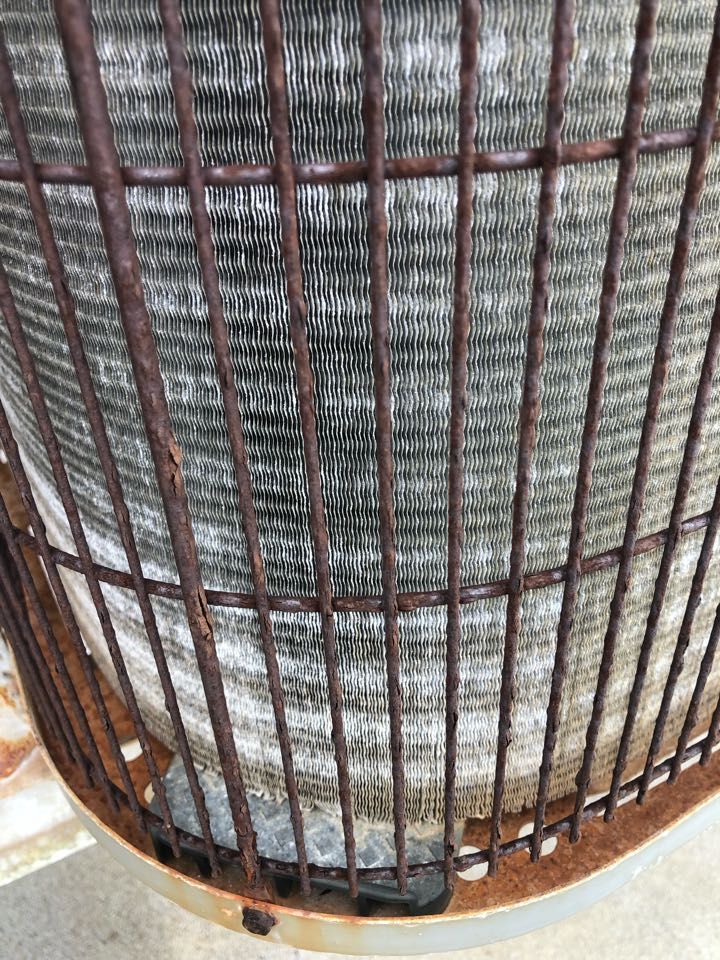 Close-up of a rusty, circular metal grate covering the metallic fins of an outdoor air conditioning unit.