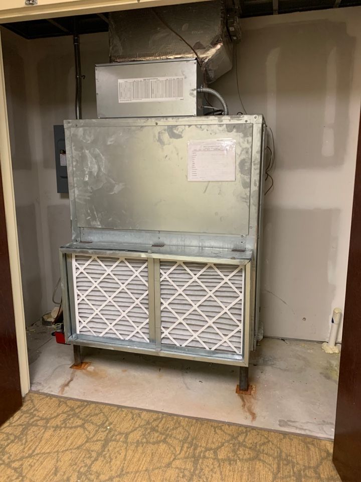 An industrial HVAC unit with two pleated filters sits inside a utility closet with light-colored walls and carpet.