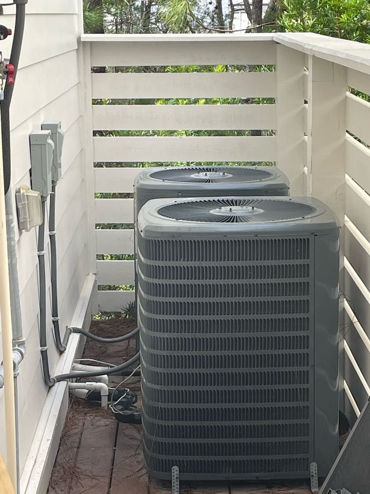 Two gray outdoor HVAC condenser units sit on a deck behind a white slatted privacy fence.