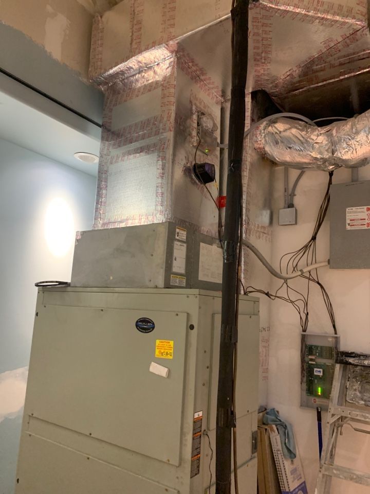 An HVAC air handler unit connected to overhead silver insulation ductwork in a utility room.
