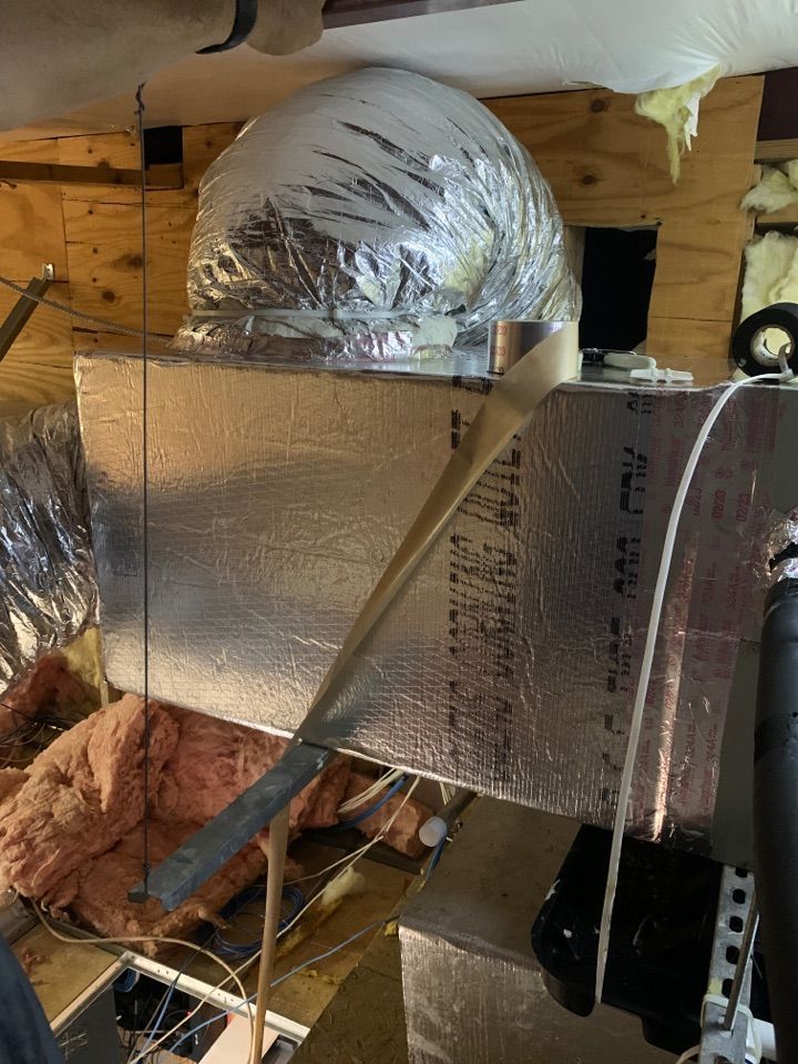 HVAC metal ductwork wrapped in silver foil insulation, suspended in an attic space with exposed wooden beams and fiberglass.