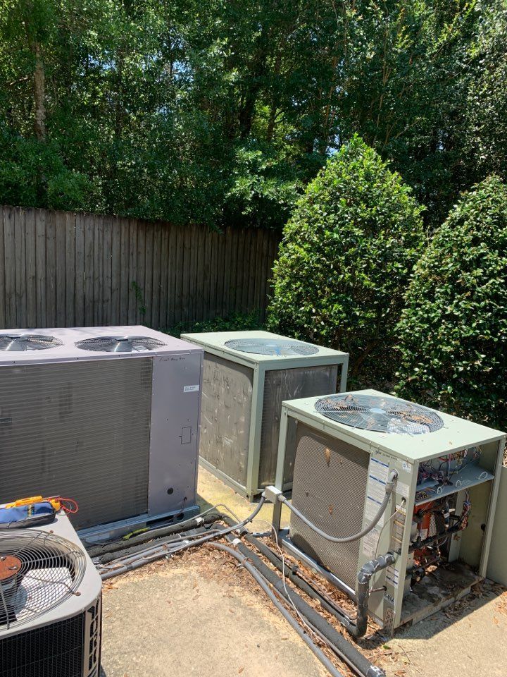 Four outdoor HVAC units placed on a concrete slab near a wooden fence and tall green bushes.