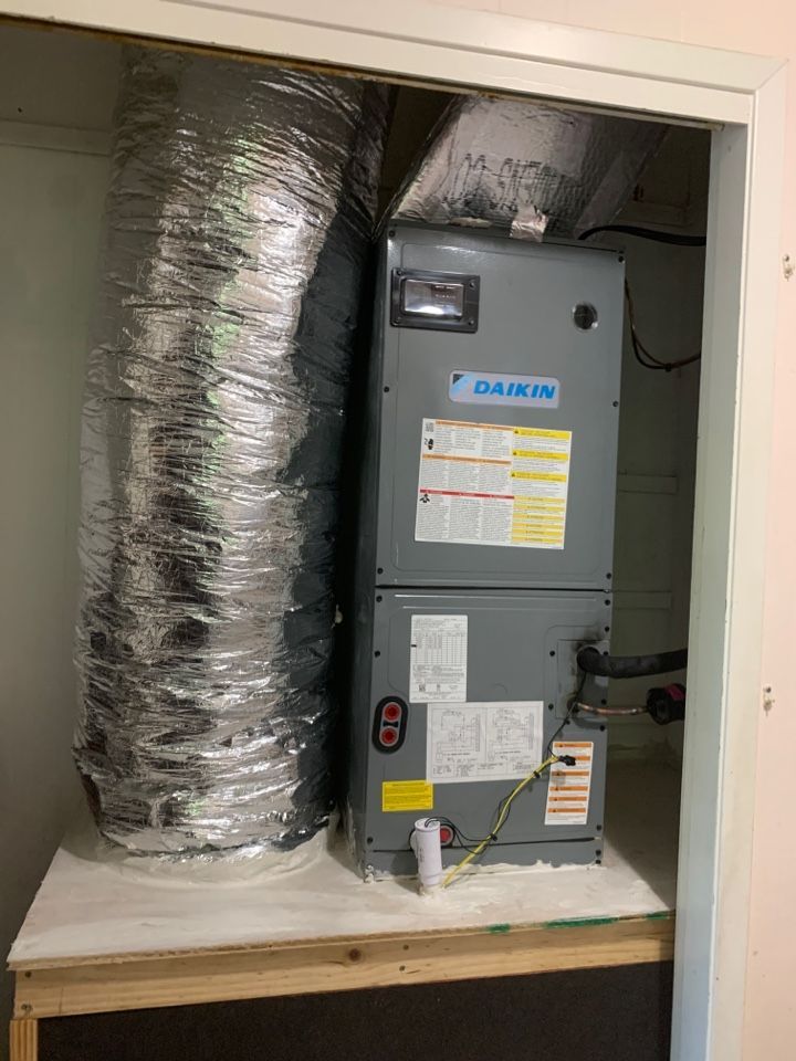 A gray Daikin HVAC air handler unit sits on a wooden platform next to a large, silver flexible duct in a closet.