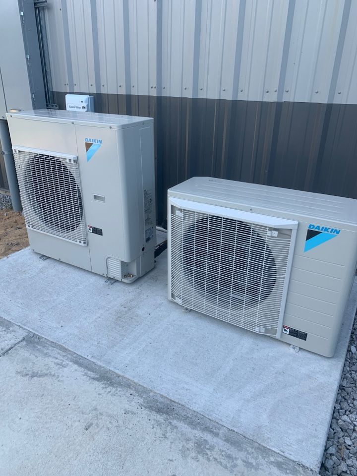 Two Daikin outdoor air conditioning units on a concrete pad against a corrugated metal wall.