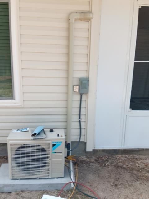 A beige Daikin outdoor HVAC unit sits on a concrete pad next to a house wall with a vertical conduit and electrical box.
