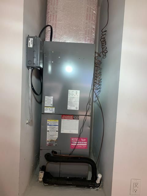 A grey HVAC air handler unit installed inside a narrow wall alcove, with black insulated piping at the base.