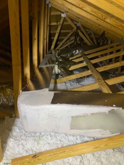 A high-angle view of an attic interior featuring wooden roof rafters, a large white duct, and loose-fill insulation.
