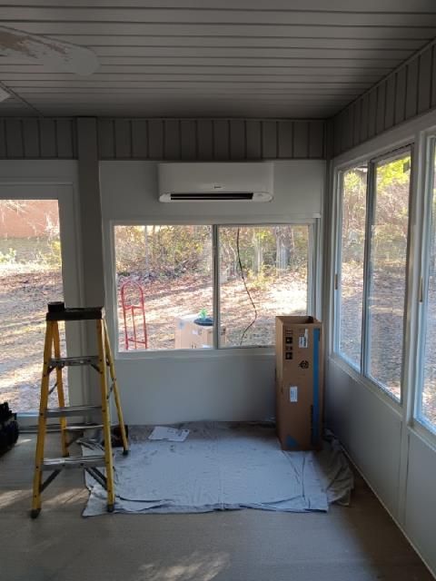 A white mini-split AC unit mounted on a wall above a window in a sunroom with a yellow stepladder and a cardboard box.