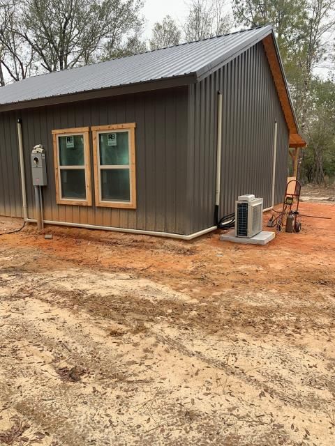 A dark brown metal-sided shed with two windows, a metal roof, and an exterior HVAC unit sits on a red dirt plot.