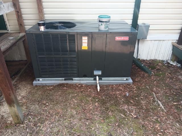 A dark gray Goodman HVAC package unit sits outdoors on the ground next to a light-colored house.