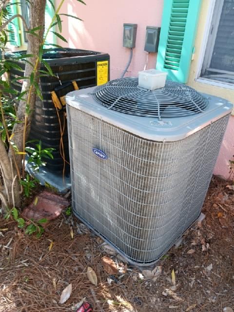 Two outdoor HVAC units situated on mulch next to a house with pink walls and green shutters.