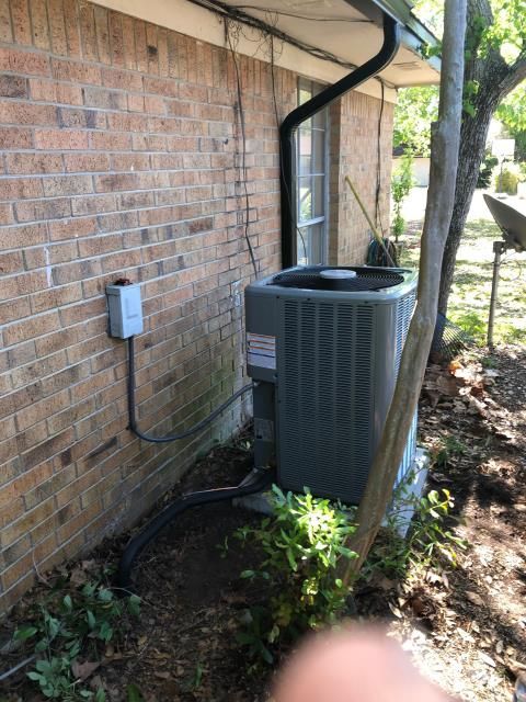 An air conditioning unit sits on a concrete pad next to a brick house exterior with a metal electrical disconnect box.
