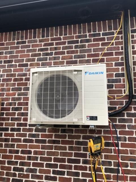 A Daikin outdoor HVAC unit mounted on a red brick exterior wall with exposed wiring and a yellow tool hanging nearby.