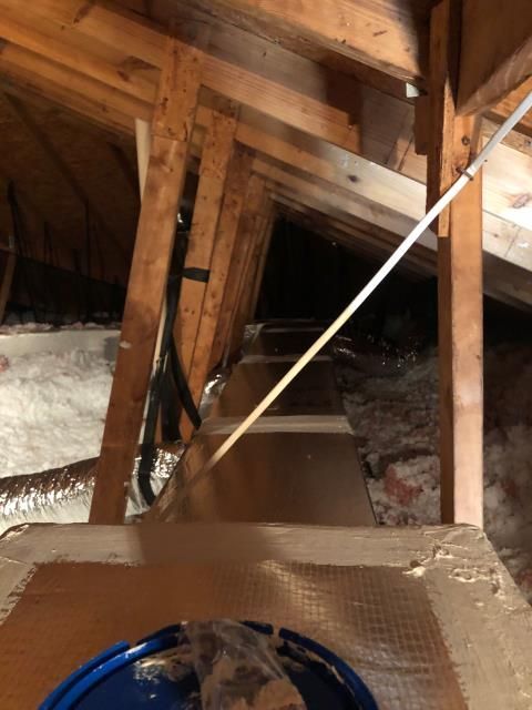 A view into a dimly lit attic showing wooden roof trusses, insulated ductwork, and loose-fill insulation between joists.
