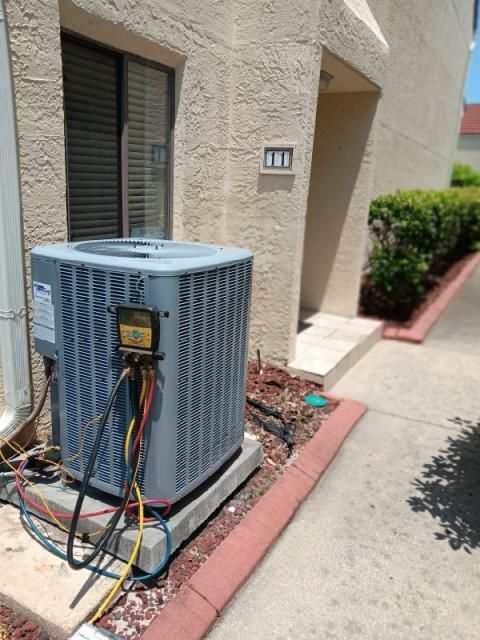 An HVAC technician's pressure gauge manifold is connected to an outdoor air conditioning unit beside a building wall.