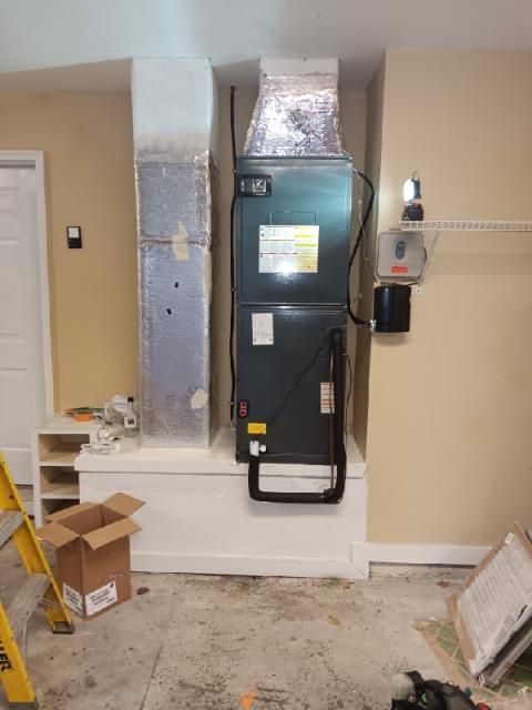 A gray HVAC air handler unit sitting on a white platform base in a room with tan walls and exposed concrete flooring.