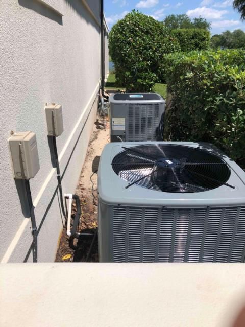 Two gray air conditioning units installed outdoors along the side of a building, next to a manicured hedge.