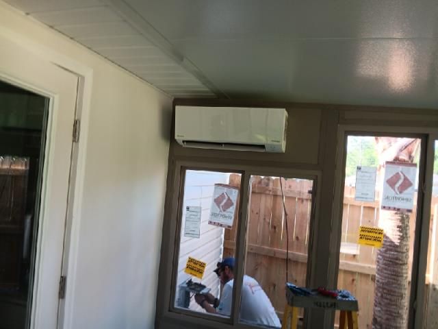 A technician works on a wall-mounted AC unit in a bright sunroom with large windows.