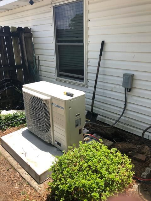 A beige outdoor HVAC condenser unit sits on a concrete pad next to a house wall with white siding, a window, and a bush.