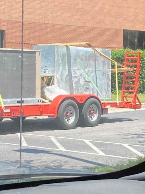 A red utility trailer parked in a lot carries a large, silver industrial HVAC unit secured with yellow straps.