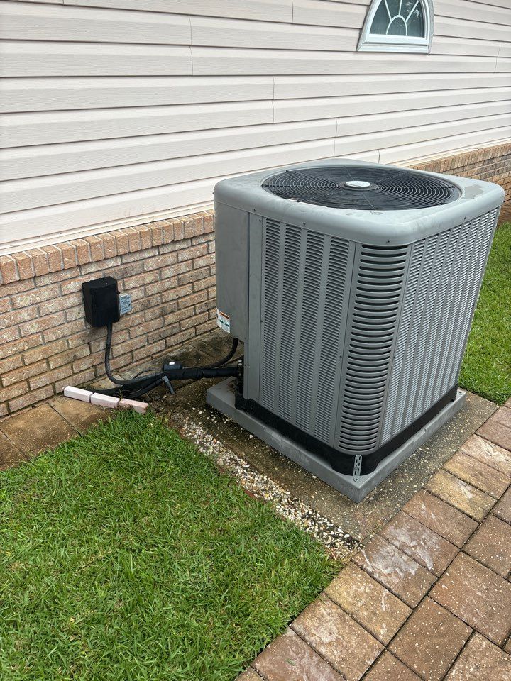 A grey HVAC condenser unit sits on a concrete pad next to a brick-and-siding exterior wall with a black electrical box.