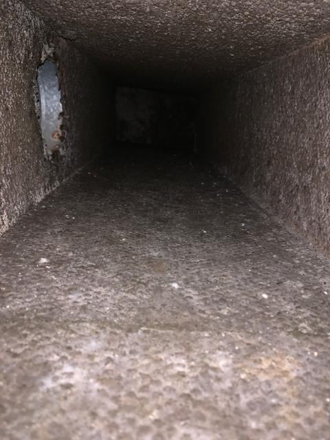 An interior view looking down a dusty, rectangular ventilation duct with a metal seam on the left wall.