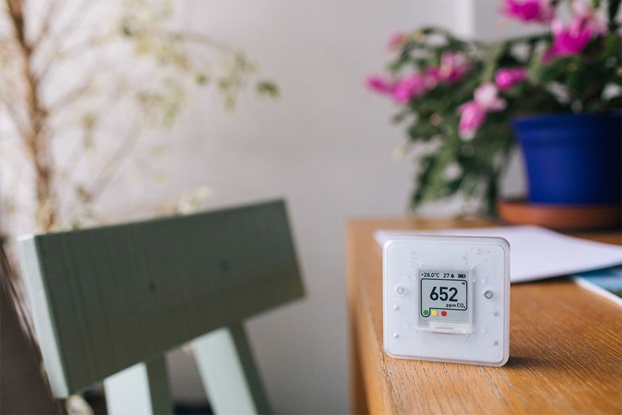 White digital sensor on a wooden table beside flowers and a chair in a bright room