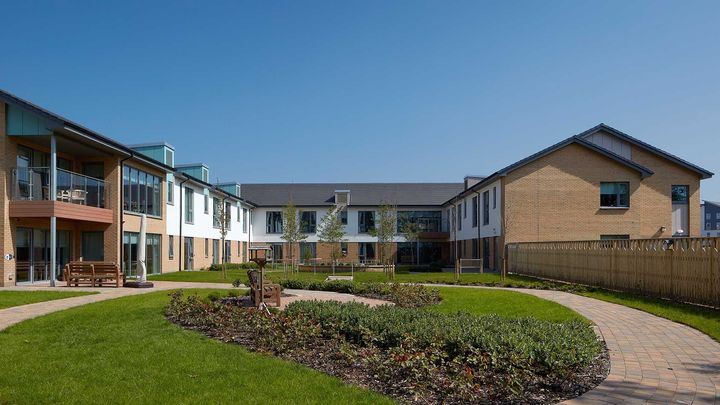 Apartment courtyard with green lawn, landscaped bushes, and walkways beside brick and white buildings under a clear sky
