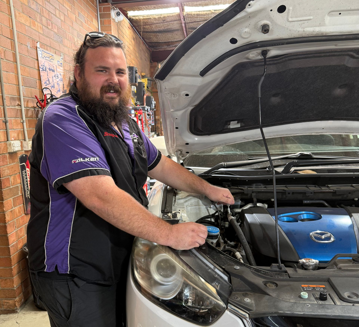 Andrew, a qualified Newcastle Mechanic smiling beside a car with the hood open in the workshop — AutoHop Lambton in Lambton, NSW