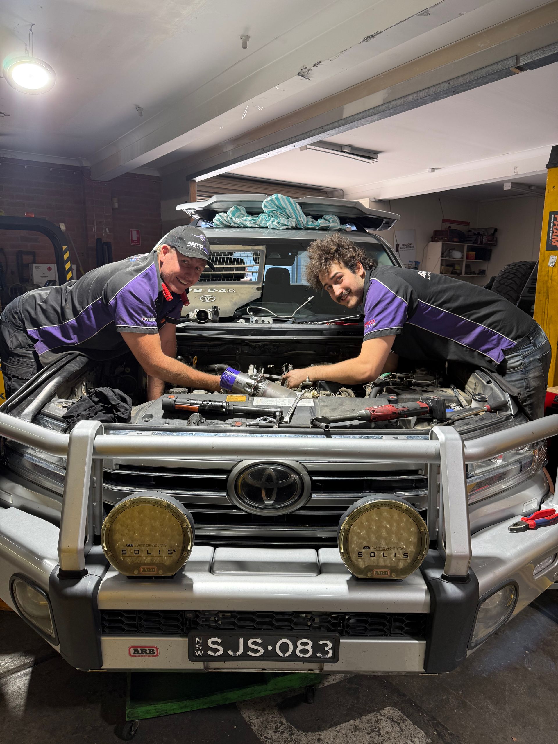 Two Newcastle mechanics in purple uniforms working on a car engine in the AutoHop Lambton workshop