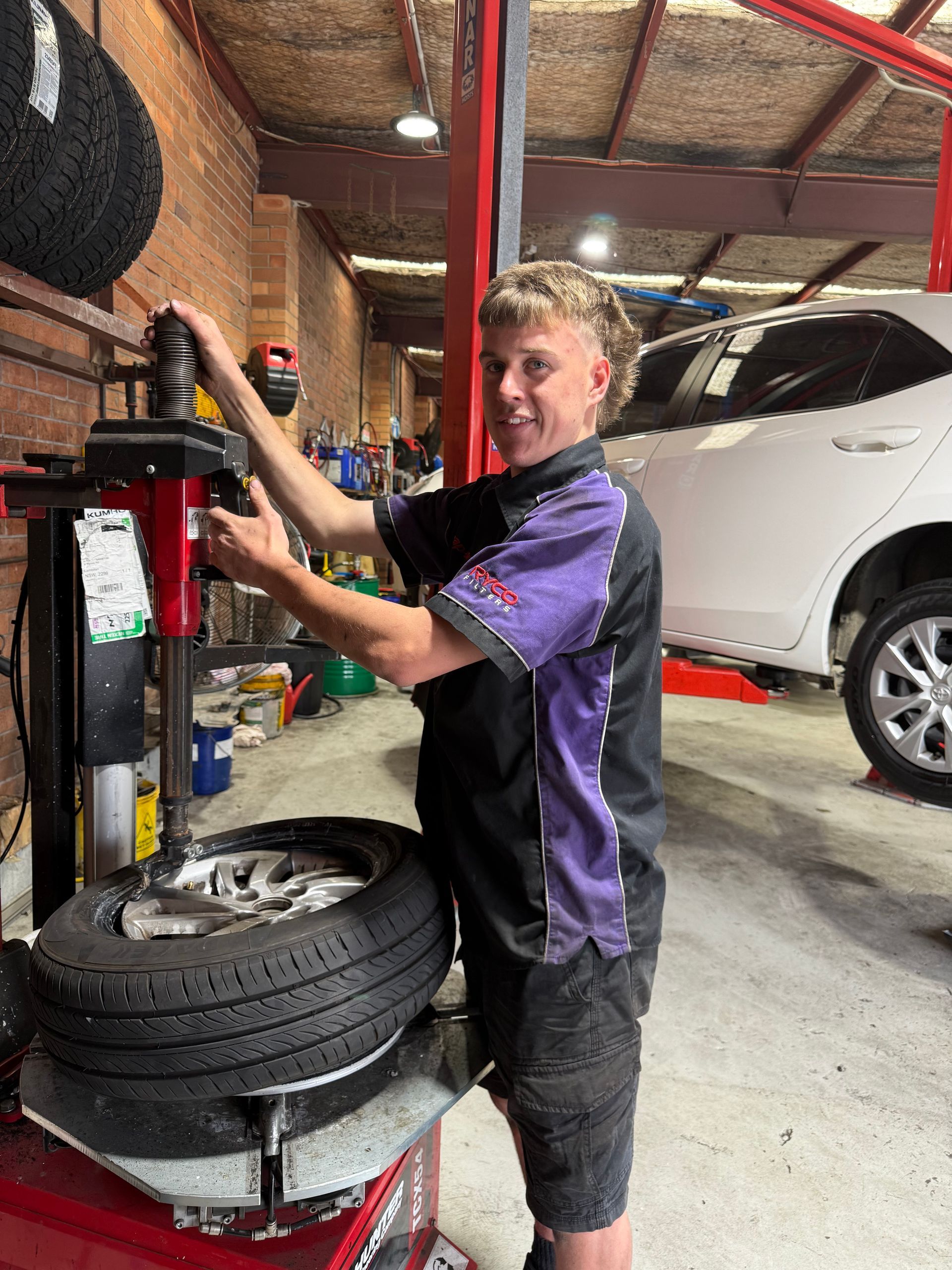 Mechanic at a tyre machine in a shop; he is smiling, wearing work clothes.— AutoHop Lambton in Lambton, NSW