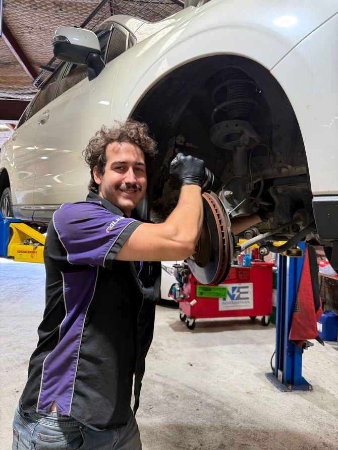 Mechanic smiling while working on a raised white car in an auto shop — AutoHop Lambton in Lambton, NSW
