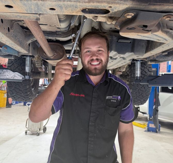Smiling mechanic under a lifted vehicle, holding a wrench in a repair shop — AutoHop Lambton in Lambton, NSW