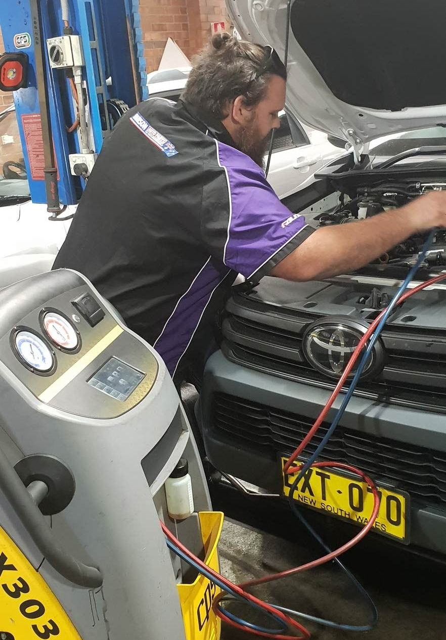 Mechanic in A Purple and Black Uniform Working on A Car with An Ac Machine in An Auto Shop — AutoHop Lambton in Lambton, NSW