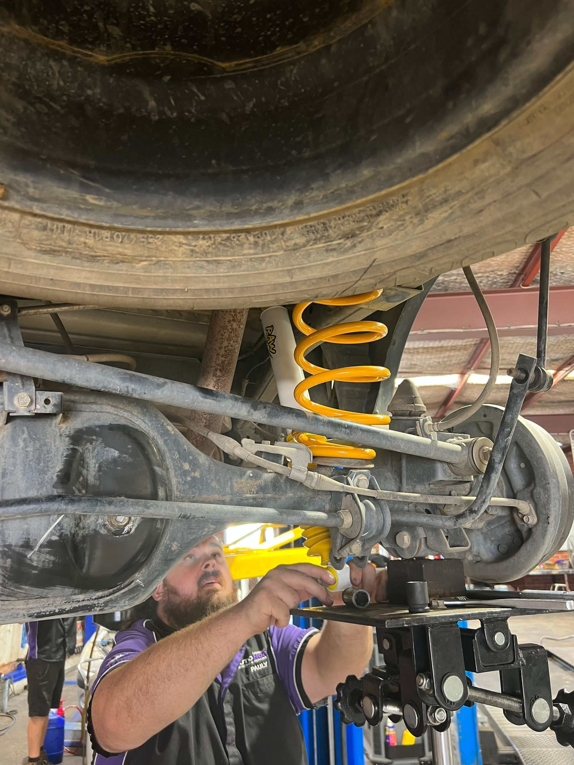 Mechanic Working on The Underside of A Vehicle with A Yellow Coil Spring — AutoHop Lambton in Lambton, NSW