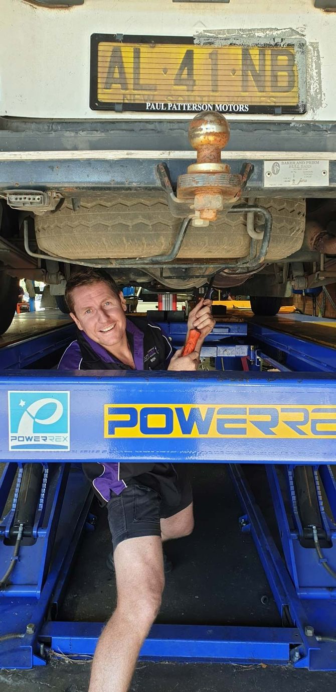 Man smiling under the rear of a blue tow truck with a yellow license plate — AutoHop Lambton in Lambton, NSW